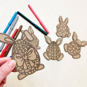 May include: Four unfinished wooden Easter bunny ornaments with intricate designs. The bunnies are holding eggs with patterns. One bunny is being held by a hand, and there are several colored markers nearby.