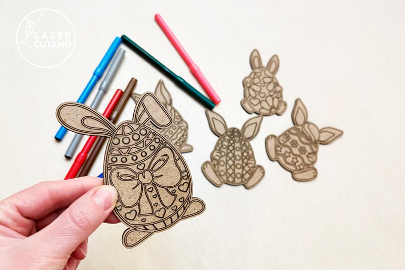 May include: Four unfinished wooden Easter bunny ornaments with intricate designs. The bunnies are holding eggs with patterns. One bunny is being held by a hand, and there are several colored markers nearby.