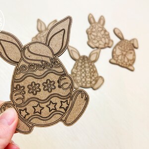 May include: Five wooden Easter bunny ornaments, each shaped like a bunny holding an egg. The eggs are decorated with different patterns, including swirls, stars, and flowers. The ornaments are a light brown color.