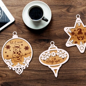 Puede incluir: Tres adornos navide&ntilde;os de madera blanca con dise&ntilde;os intrincados. Un adorno presenta un mu&ntilde;eco de nieve con el texto "let it snow", otro tiene el texto "Holy night", y el tercero es una estrella con un mu&ntilde;eco de nieve dentro.