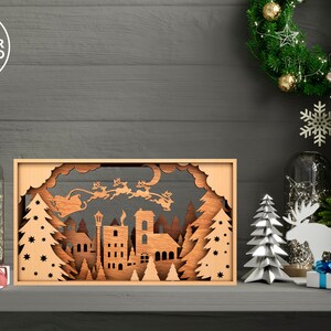 May include: A wooden Christmas scene with a cutout of Santa Claus flying in a sleigh pulled by reindeer over a town with trees and stars. The scene is illuminated with lights and is surrounded by other Christmas decorations.