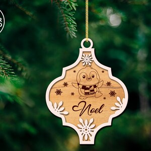 Puede incluir: Un adorno navide&ntilde;o de madera con un contorno blanco y un ping&uuml;ino con una bufanda. El adorno dice "No&euml;l" en el centro.