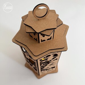 May include: A decorative wooden lantern with a star-shaped top and intricate cut-out designs. The lantern is made of light brown wood and has a small handle at the top. The Laser Cutano logo is in the upper left corner.