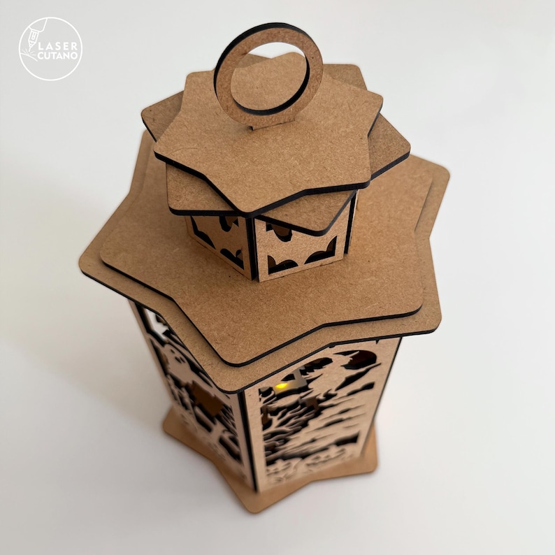 May include: A decorative wooden lantern with a star-shaped top and intricate cut-out designs. The lantern is made of light brown wood and has a small handle at the top. The Laser Cutano logo is in the upper left corner.