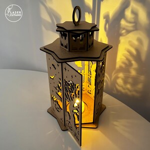 May include: A wooden Halloween lantern with intricate cut-out designs of bats, trees, and pumpkins. The lantern has a handle and a door that opens to reveal a lit candle inside. The Laser Cutano logo is visible in the upper left corner.