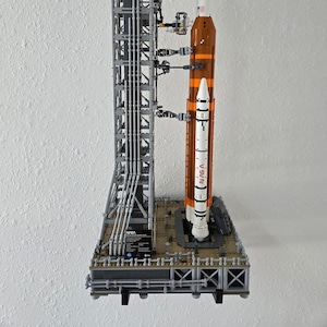 May include: A detailed LEGO model of a NASA Space Launch System rocket on a launchpad. The rocket is white and orange with the NASA logo and "SLS" printed on the side. The launchpad is gray and black with a detailed structure and support beams.