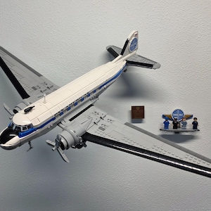 May include: A model airplane, constructed from interlocking plastic bricks, is predominantly white with blue and gray accents. The words "PAN AMERICAN" are visible on the fuselage. A small display features miniature figures and a logo.