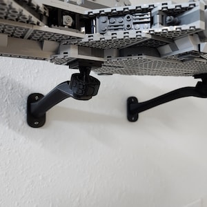 May include: Two black wall-mounted brackets supporting a large gray plastic building block model of a spaceship. The brackets feature a curved arm design and a circular mounting base. One bracket displays the embossed text P3D on its connector.