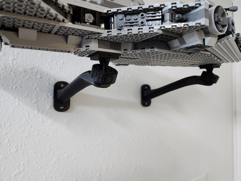 May include: Two black wall-mounted brackets supporting a large gray plastic building block model of a spaceship. The brackets feature a curved arm design and a circular mounting base. One bracket displays the embossed text P3D on its connector.