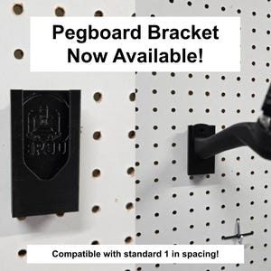 May include: A black pegboard bracket mounted on a white pegboard with holes. The text reads Pegboard Bracket Now Available! and Compatible with standard 1 in spacing! The bracket features a shield-shaped logo with the text 3R30 on its front surface.