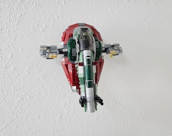 May include: A green and red Lego model of a spaceship, the Slave I, from the Star Wars franchise. The model is mounted on a white wall.