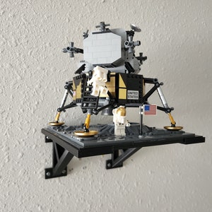 May include: A Lego model of the Apollo 11 Lunar Module, with an astronaut figure and an American flag, mounted on a black shelf.