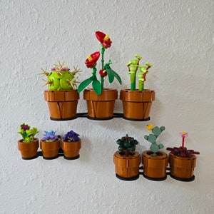 May include: A set of eight miniature potted plants made of Lego bricks. The plants are arranged in a row on a white wall. The plants include a cactus, a succulent, a fern, a flower, and a vine. The pots are all brown and have a simple design.