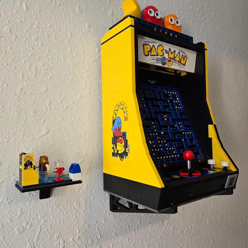 May include: A yellow and black Lego Pac-Man arcade game with a blue and white maze and red joystick. The game is mounted on a black wall mount. The game is a replica of the classic arcade game.