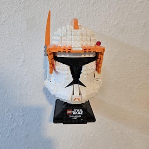 May include: LEGO Star Wars Commander Cody helmet build. The helmet is white with orange accents and a black visor. The base is black with the LEGO Star Wars logo and the text "Commander Cody".