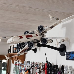 May include: A white and dark red plastic building block model of an X-wing starfighter mounted on a black wall stand, positioned above a shelf containing numerous Star Wars action figures including stormtroopers and various armored characters.