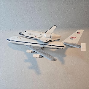 Pode incluir: Um modelo Lego branco de um vaivém espacial da NASA montado em cima de um modelo Lego branco de um Boeing 747. O 747 tem o logótipo da NASA e o número 905 na cauda. O vaivém espacial tem as palavras "United States" na lateral.
