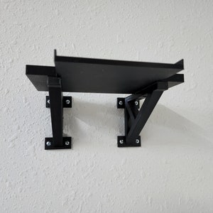 May include: Black shelf with two triangular metal brackets attached to a white wall. The shelf is made of wood and has a simple design.