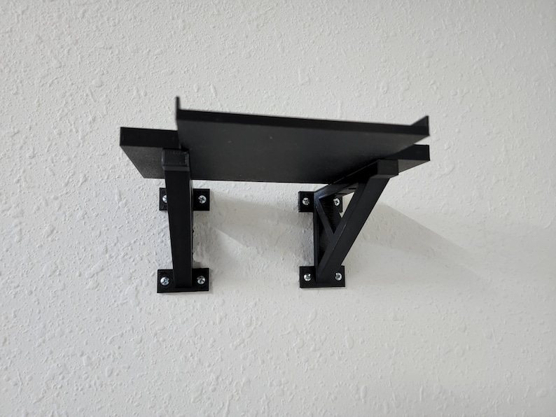 May include: Black shelf with two triangular metal brackets attached to a white wall. The shelf is made of wood and has a simple design.