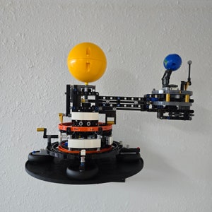 May include: A black and silver LEGO model of a solar system with a yellow sun, a blue planet, and gears. The model is mounted on a black base.