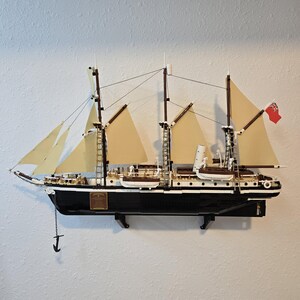 May include: A detailed model of a sailing ship with white sails and a black hull. The ship is mounted on a wall with a black stand. The ship has a red flag with a white symbol on it.