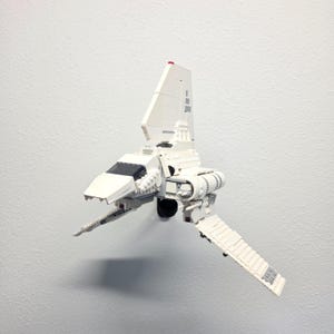 Pode incluir: Um modelo de nave espacial construído com blocos de plástico branco, apresentando uma barbatana estabilizadora vertical alta, um cockpit pontiagudo com para-brisas preto e duas grandes asas articuladas que se estendem para baixo a partir do corpo.