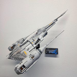 May include: A detailed grey and yellow starfighter model, with multiple engines and a long, pointed front. The model is mounted on a white wall, with a small information card visible.