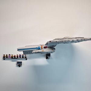 May include: A large, gray Lego model of a spaceship, mounted on a white wall. The model has intricate details and is accompanied by a shelf with Lego figurines. The ship has blue and orange accents.