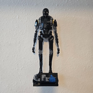 May include: A black and gray Lego K-2SO droid figure from Star Wars, standing on a black base. The figure has a rounded head, articulated limbs, and yellow accents. The base features character details and a blue schematic.