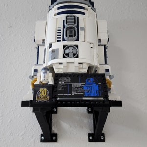 May include: A white and blue Lego R2-D2 model on a black display stand. The stand has a black plaque with the text "STAR WARS R2-D2" and a small Lego R2-D2 figure. The plaque also includes information about the model, including its manufacturer, function, and processing indicator color.