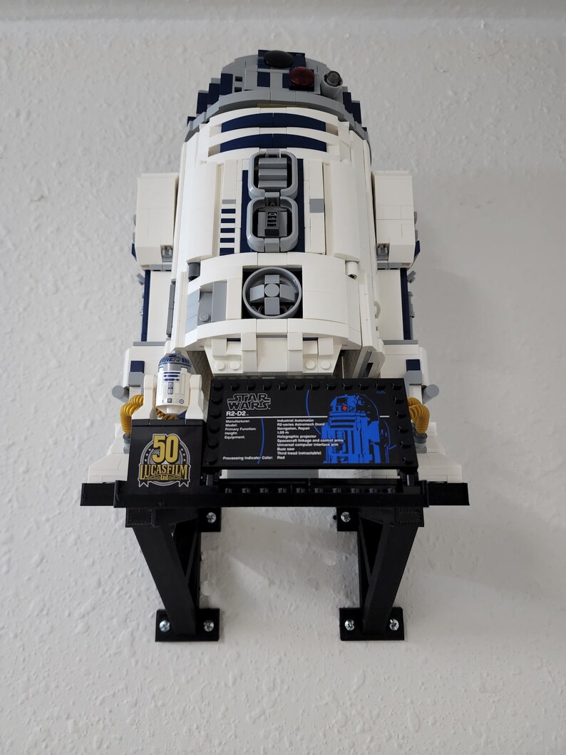 May include: A white and blue Lego R2-D2 model on a black display stand. The stand has a black plaque with the text "STAR WARS R2-D2" and a small Lego R2-D2 figure. The plaque also includes information about the model, including its manufacturer, function, and processing indicator color.