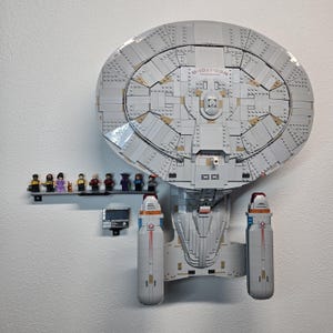 May include: A large, gray Lego model of a spaceship, mounted on a white wall. The model has a circular main body and two cylindrical engines. A small shelf holds several Lego figures. The model is labeled "NCC-1701-D".