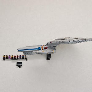 May include: A large gray plastic building block model of a starship mounted on a wall. Below the ship, a small black display shelf holds nine miniature figures, and a black rectangular plaque with white text is mounted to the wall.