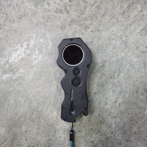 May include: A black handheld device with a circular lens and two smaller circular buttons. The device has a textured surface and a lanyard attached to the bottom. The background is a gray concrete surface.