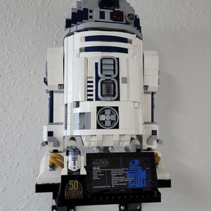 May include: A white and blue LEGO Star Wars R2-D2 model with a black display stand. The stand features a small LEGO R2-D2 figure and a black plaque with the text "STAR WARS R2-D2" and technical specifications. The stand also has a 50th anniversary Lucasfilm logo.
