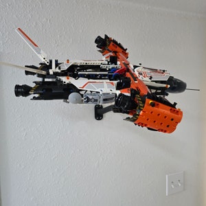 May include: A white, orange, and black Lego spaceship model with a detailed design. The spaceship is mounted on a black wall mount.