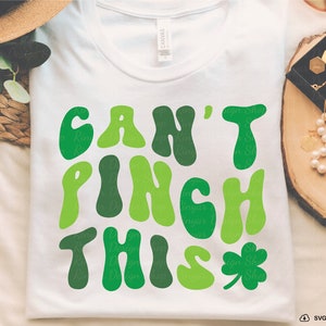 Peut inclure: Un t-shirt blanc avec un graphique vert qui dit "Can't Pinch This" dans une police r&eacute;tro. Une tr&egrave;fle vert se trouve &agrave; droite du texte.
