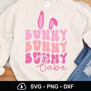 Może przedstawiać: Kremowa bluza z różowymi uszami królika i napisem "Bunny Bunny Bunny Babe" w odcieniach różu. Projekt ma czcionkę retro. Bluza jest połączona z niebieskimi dżinsami.