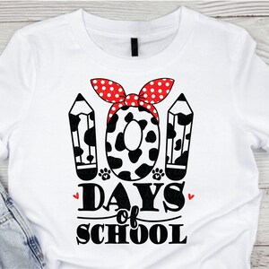 May include: White t-shirt with a black and white graphic of two pencils and a cow print circle with a red and white polka dot bow. The text "100 Days of School" is below the graphic.