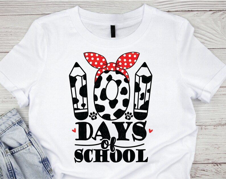 May include: White t-shirt with a black and white graphic of two pencils and a cow print circle with a red and white polka dot bow. The text "100 Days of School" is below the graphic.