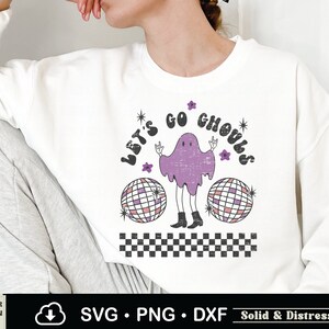 May include: A white sweatshirt with a purple ghost wearing boots and holding up peace signs. The ghost is surrounded by two disco balls and the text "Let's Go Ghouls".