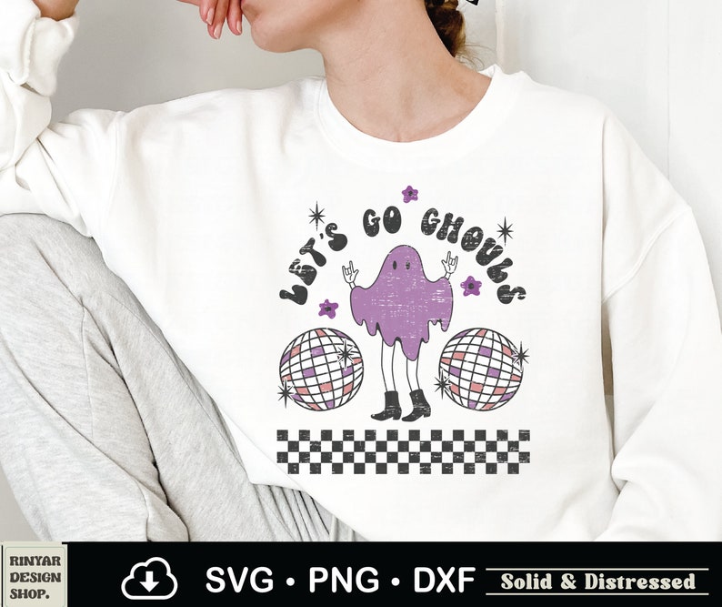 May include: A white sweatshirt with a purple ghost wearing boots and holding up peace signs. The ghost is surrounded by two disco balls and the text "Let's Go Ghouls".
