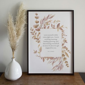 Può includere: Una stampa incorniciata con un design floreale in tonalità rosa e marrone. La stampa presenta una citazione sull'amore per se stessi e la crescita, attribuita a Idil Ahmed.