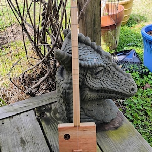 May include: A handmade wooden cigar box guitar with a carved face on the neck. The instrument has a square box body and a single string. It rests on a weathered wooden surface, with a dragon statue in the background.