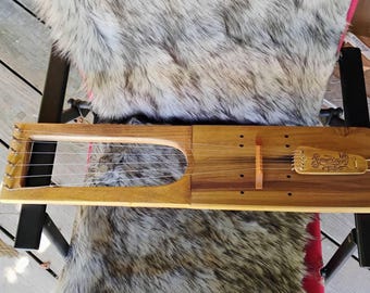 Może przedstawiać: Drewniany instrument muzyczny typu lira z sześcioma strunami rozciągniętymi na otwartej ramie i litym korpusie. Instrument posiada mostek oraz strunociąg z wygrawerowanym ozdobnym wzorem. Drewno ma naturalne, jasnobrązowe wykończenie.