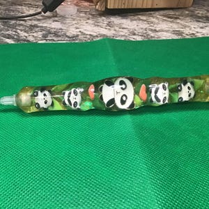 May include: A clear, resin-based diamond painting pen with a panda bear design. The pen features a gold-colored metal tip and a clear, plastic tip. The panda bears are black and white with green leaves.