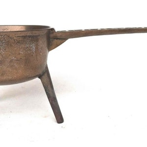 May include: A small, three-legged, bronze pot with a long handle. The pot has a worn, aged appearance.