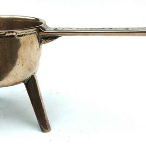 May include: A small, antique, brass pot with three legs and a long handle. The pot is made of a shiny, golden brass and has a rounded shape.