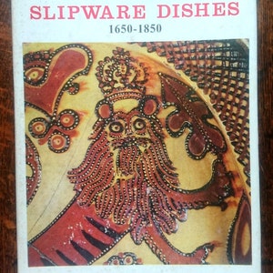 May include: Book cover with the title "English Slipware Dishes 1650-1850" and an illustration of a red and yellow lion with a crown on its head. The author is Ronald G. Cooper.