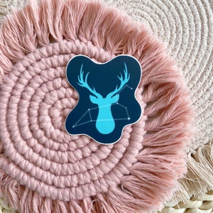 Könnte beinhalten: Eine blaue Hirsch-Silhouette mit einem Sternbild-Design auf dunkelblauem Hintergrund. Der Aufkleber befindet sich auf einer rosa und weißen gehäkelten Matte.